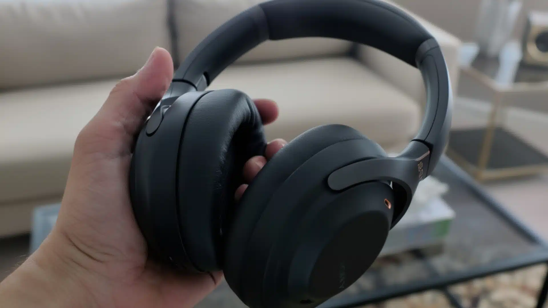 Hand holding black Sony XM3 headphones with cushioned ear cups in a living room setting