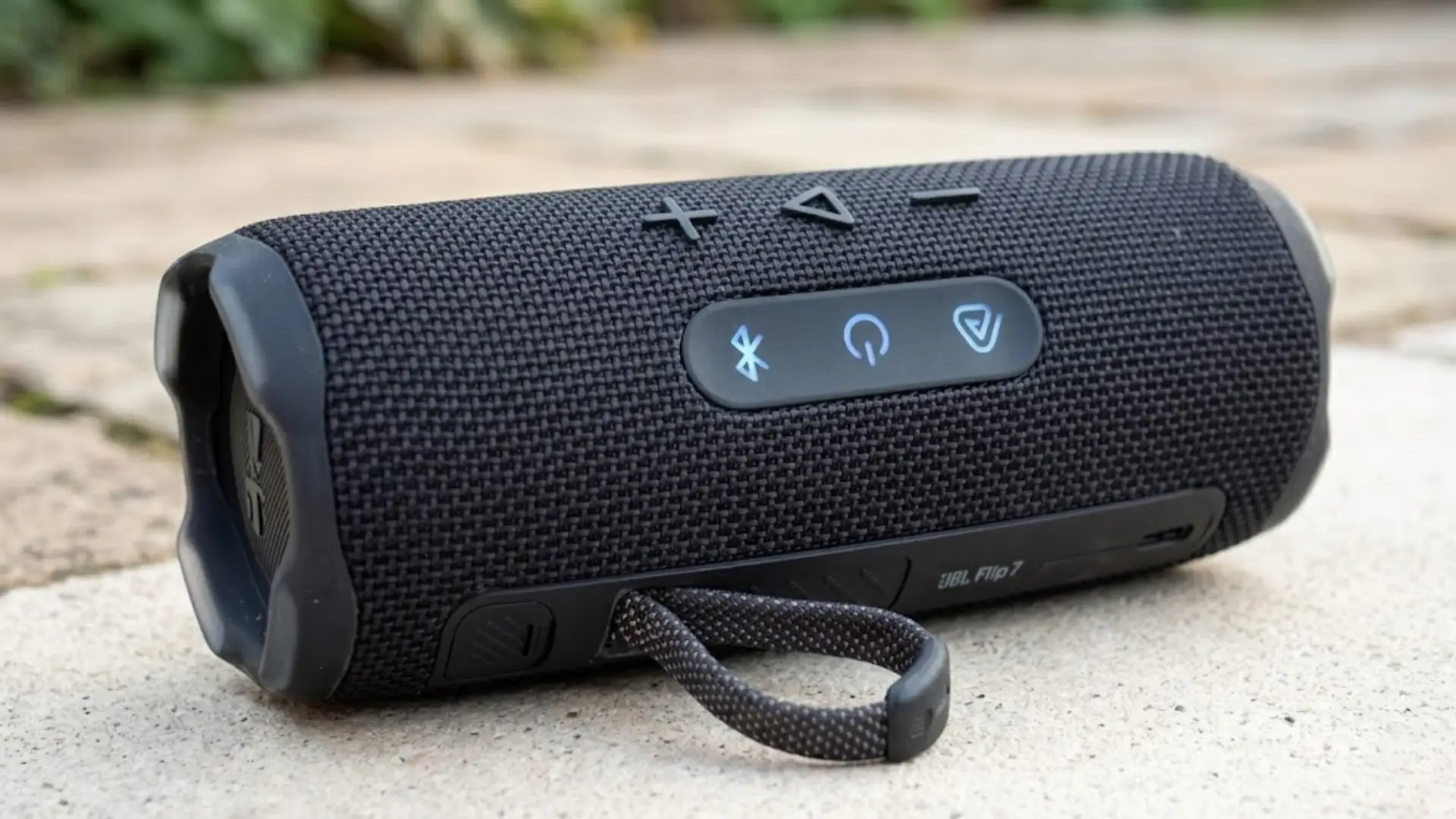 JBL Flip 7 portable speaker on outdoor surface with control buttons and strap, rugged design