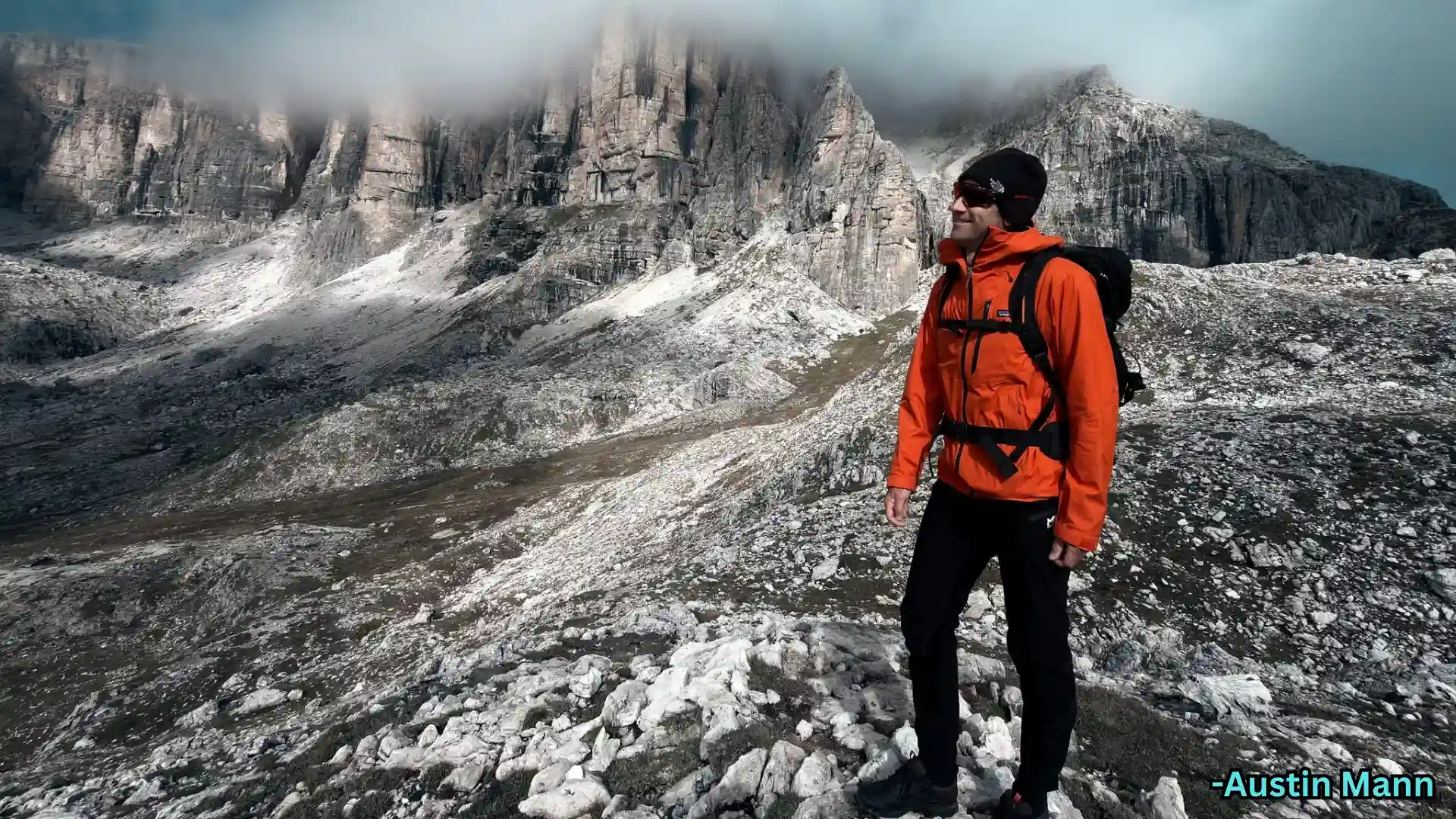 Man standing on rocky mountain terrain with backpack and orange jacket, captured on iPhone 17 with detailed landscape