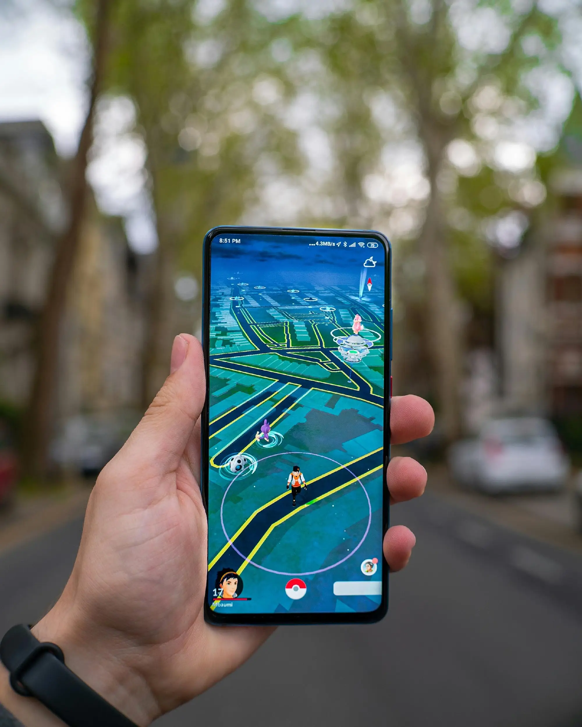 Hand holding smartphone displaying augmented reality game app on street with blurred trees in background
