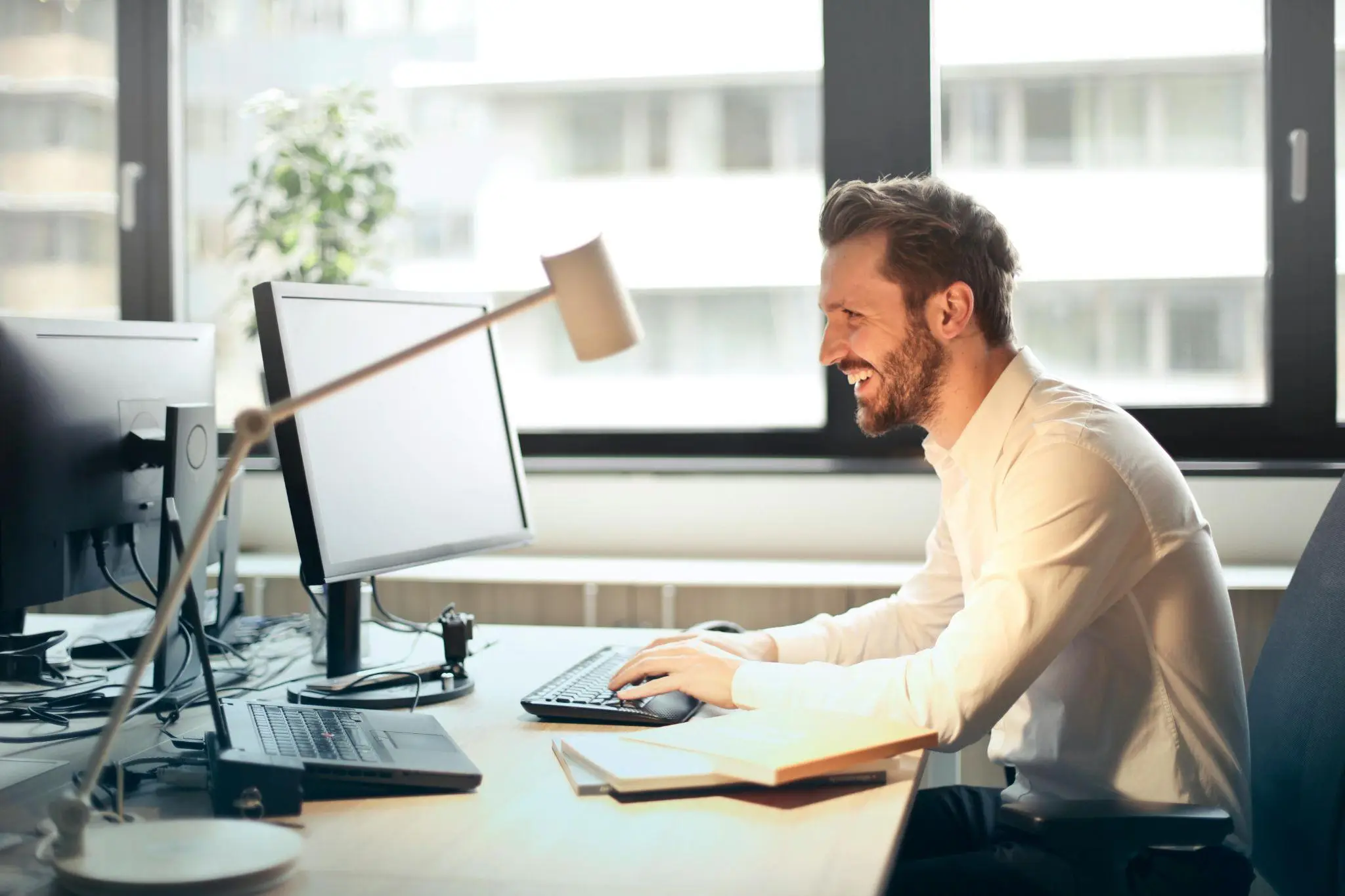 Smiling man working at a desk with computer monitors in a bright office environment