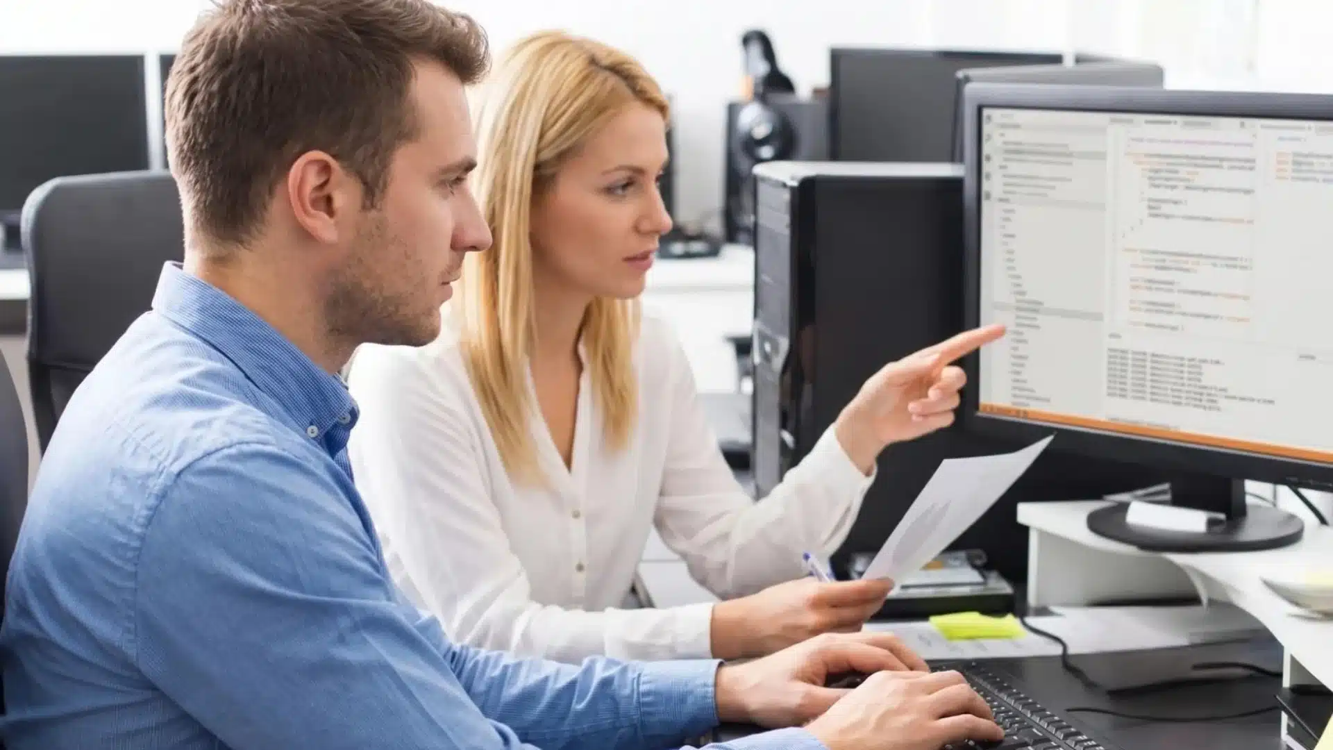 Two coworkers reviewing code on a computer, woman pointing at screen while holding a document