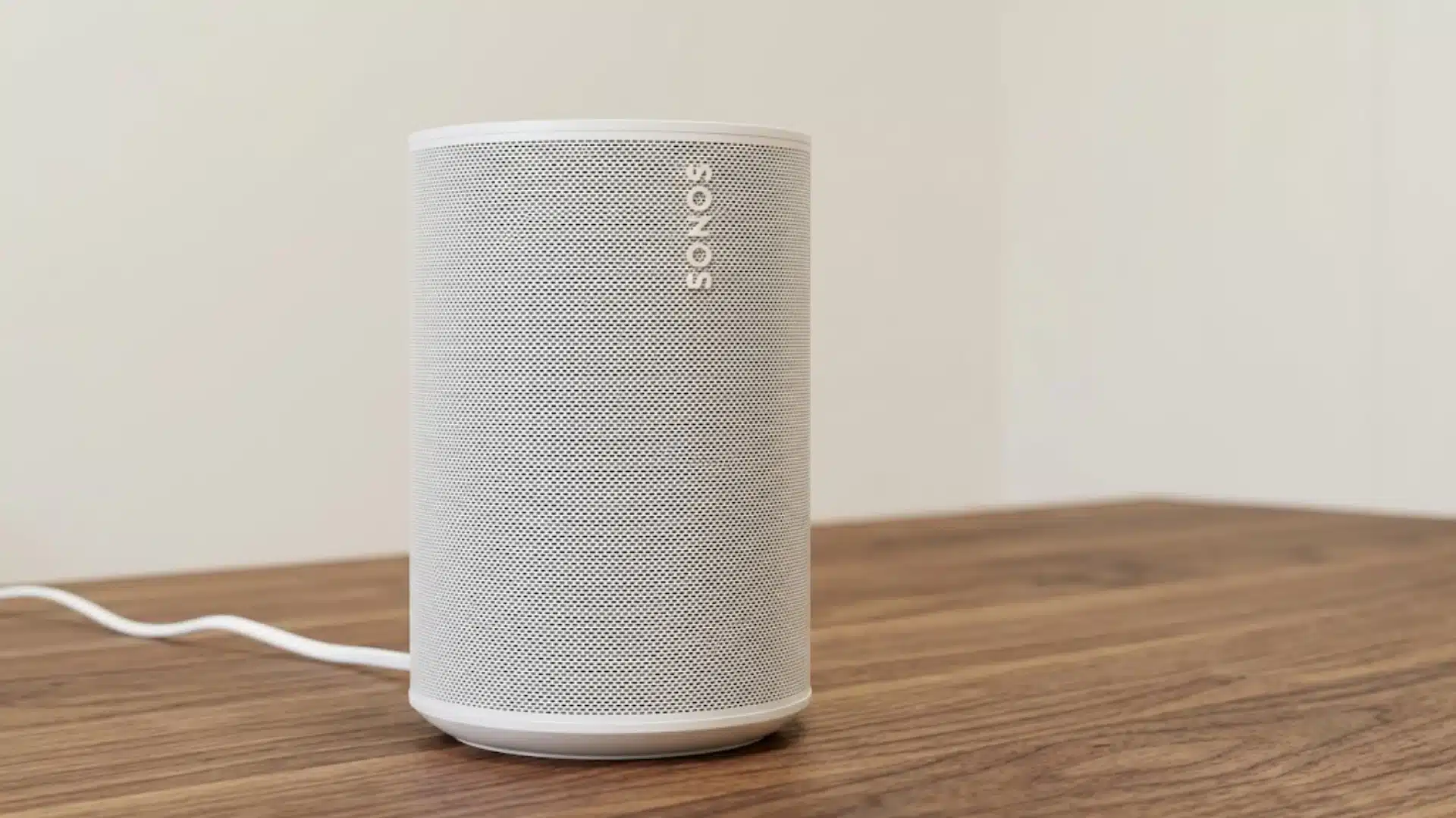 White Sonos Era 100 smart speaker on a wooden table with a power cable in a minimal home setup