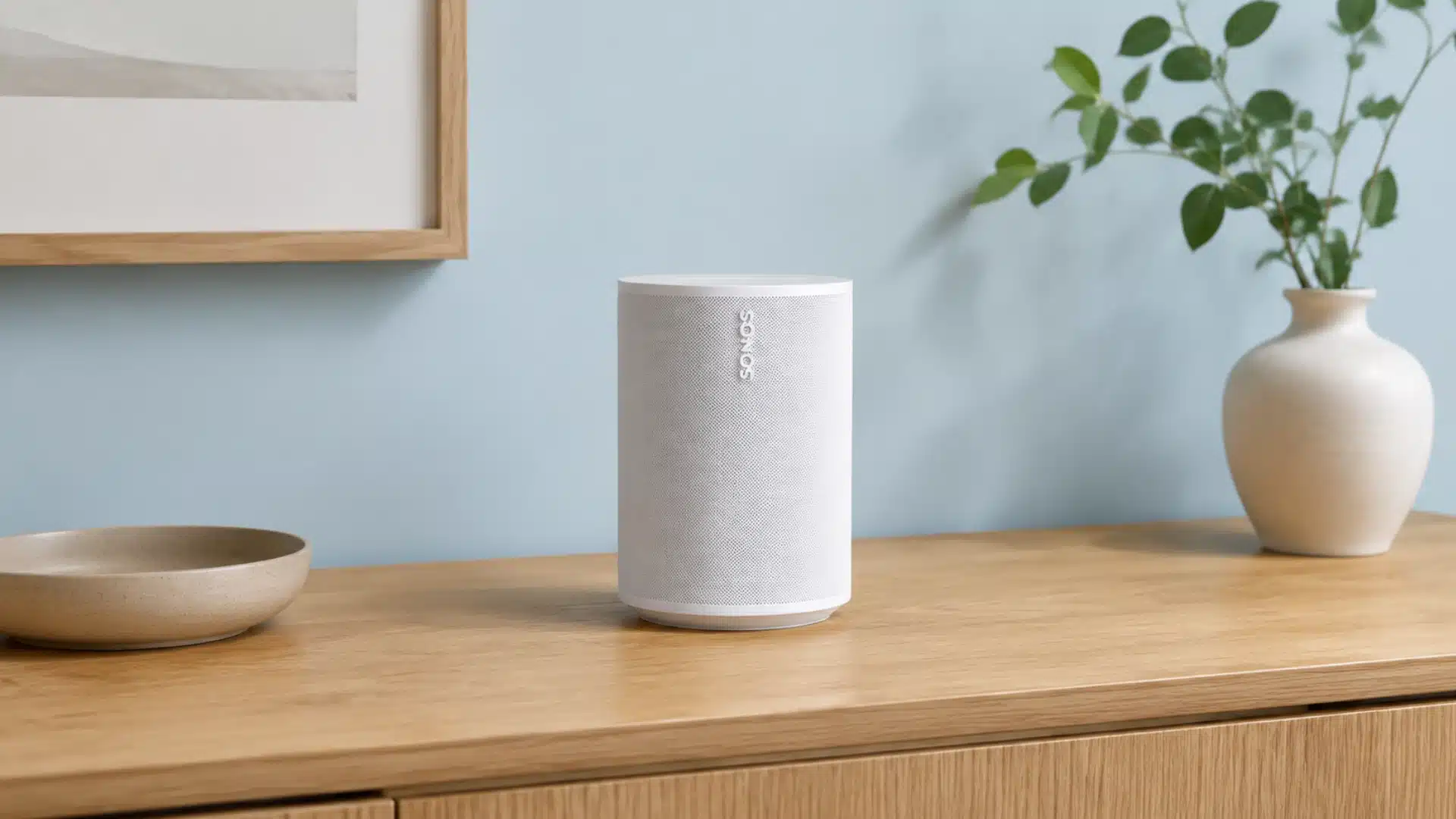 White Sonos speaker on a wooden tabletop with a light blue wall, a vase with greenery, and a minimalist ceramic bowl