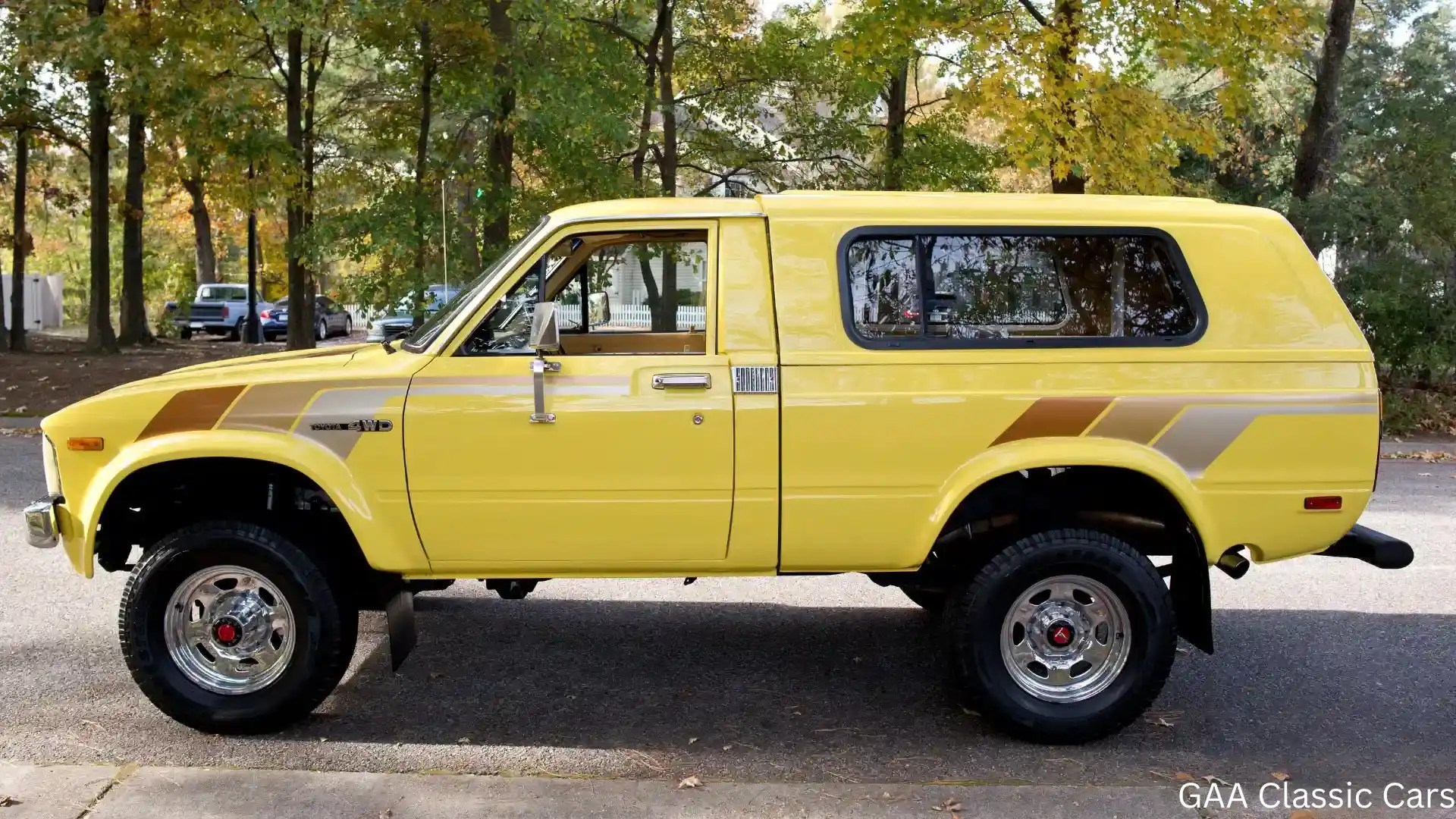 Yellow classic Toyota 4WD wagon parked beside a sidewalk with autumn trees and polished chrome wheels