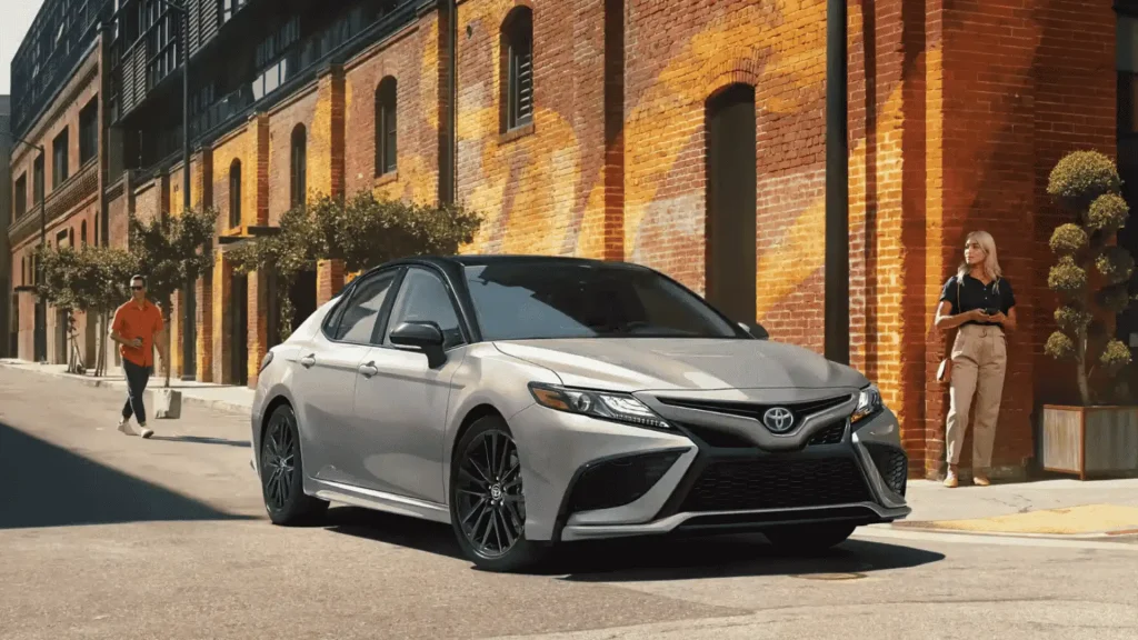 2024 Toyota Camry Hybrid sedan parked on a city street with brick buildings and pedestrians in the background