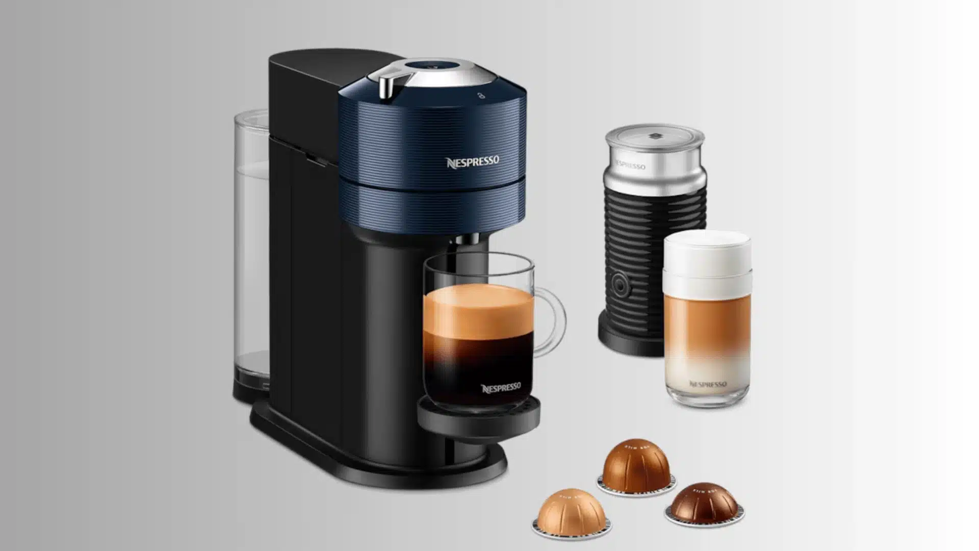 Nespresso Vertuo Next machine with coffee pods, black milk frother, and two prepared coffee drinks on a gray background