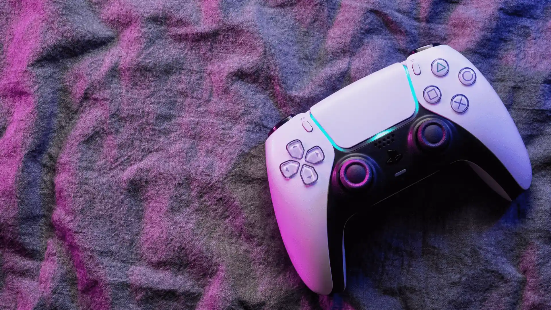 White PS5 DualSense controller illuminated by pink and blue neon lighting on a textured fabric surface