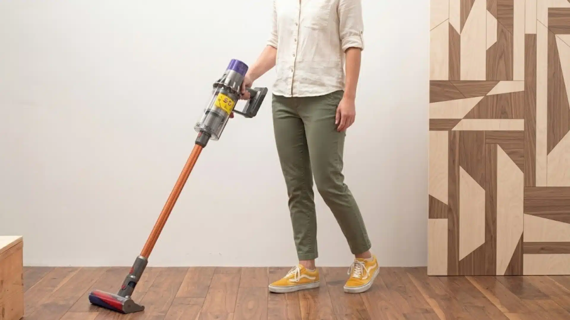 Woman using a cordless stick vacuum on hardwood flooring inside a modern home with geometric wood wall paneling