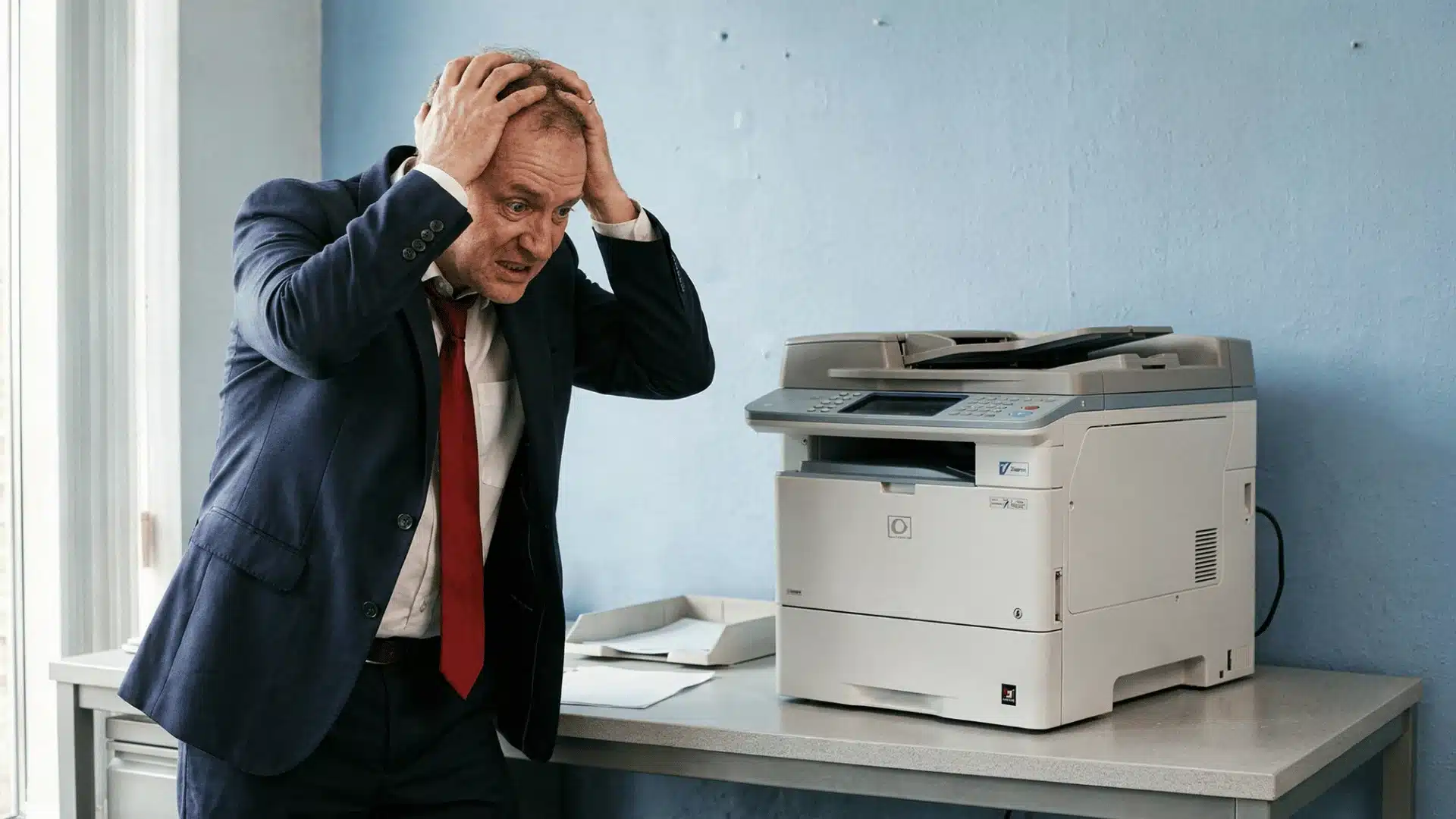 A frustrated businessman in a suit holds his head in anger next to a malfunctioning office printer