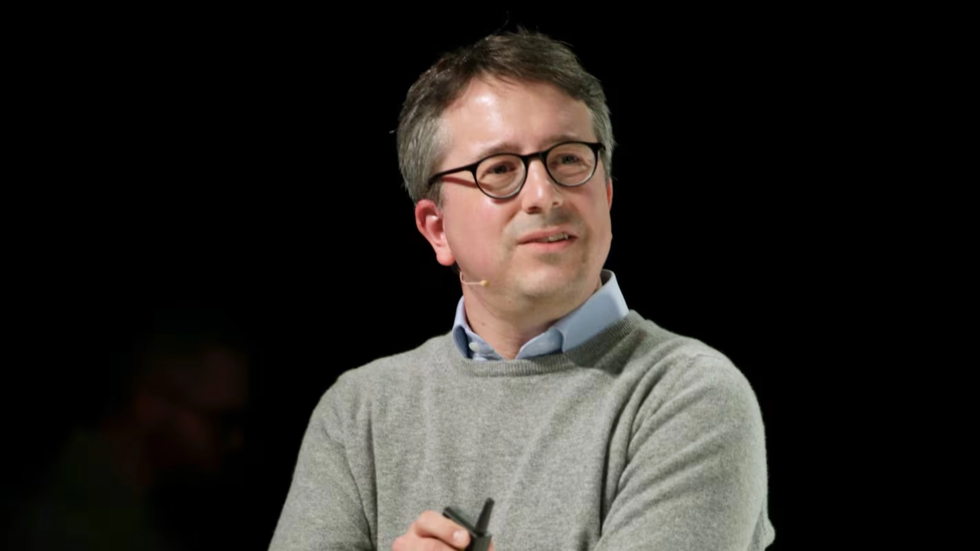 Benedict Evans in glasses holding a microphone on a dark stage background