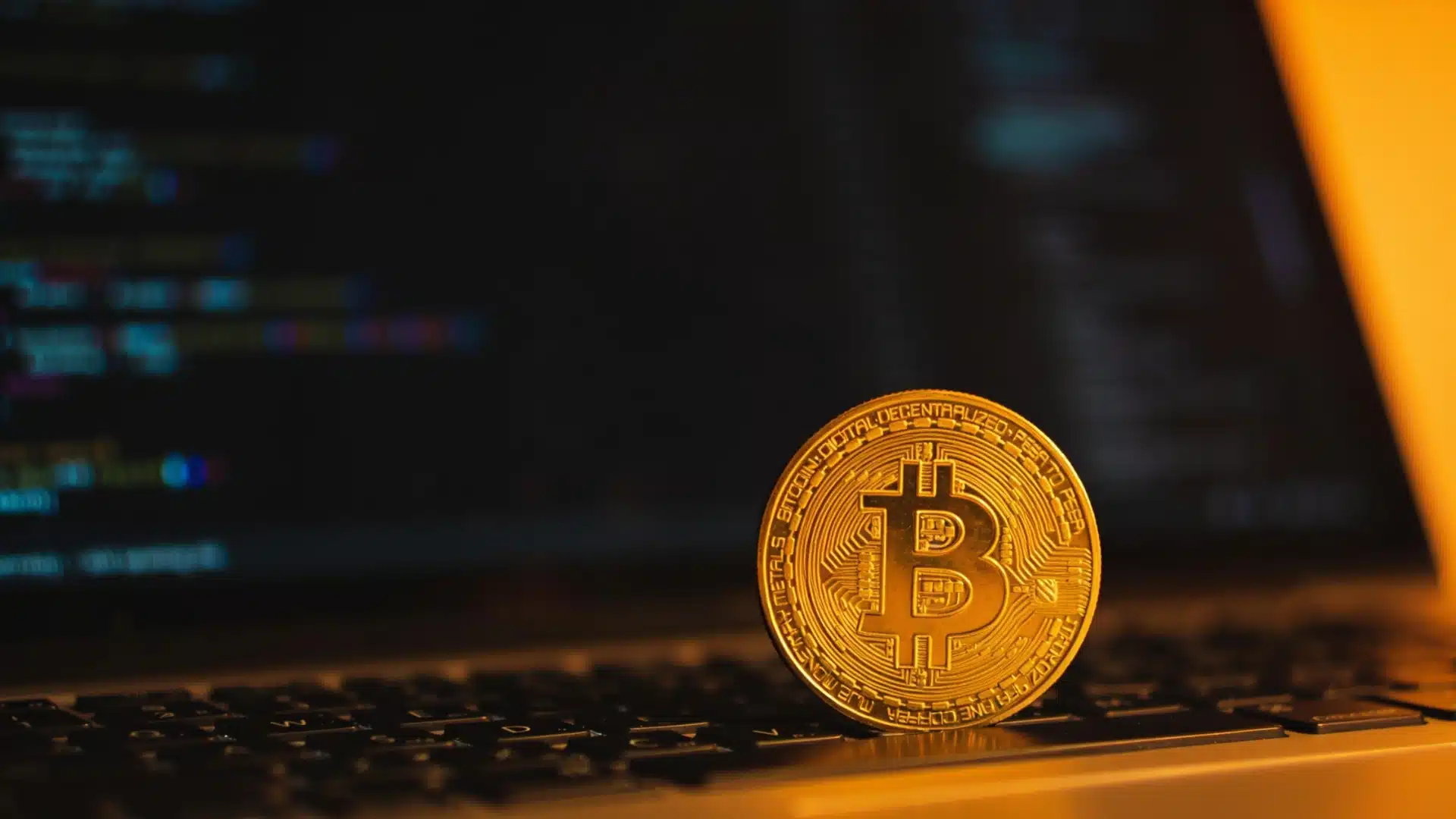 Bitcoin coin on laptop keyboard with blurred coding screen in warm light