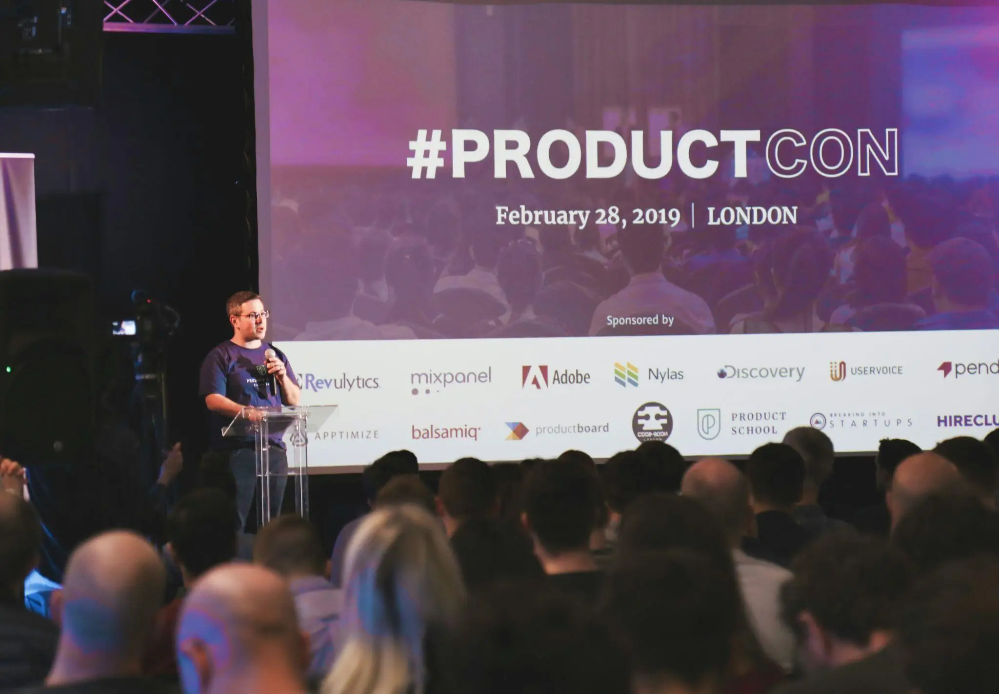 Speaker presenting at a conference with audience and large screen backdrop