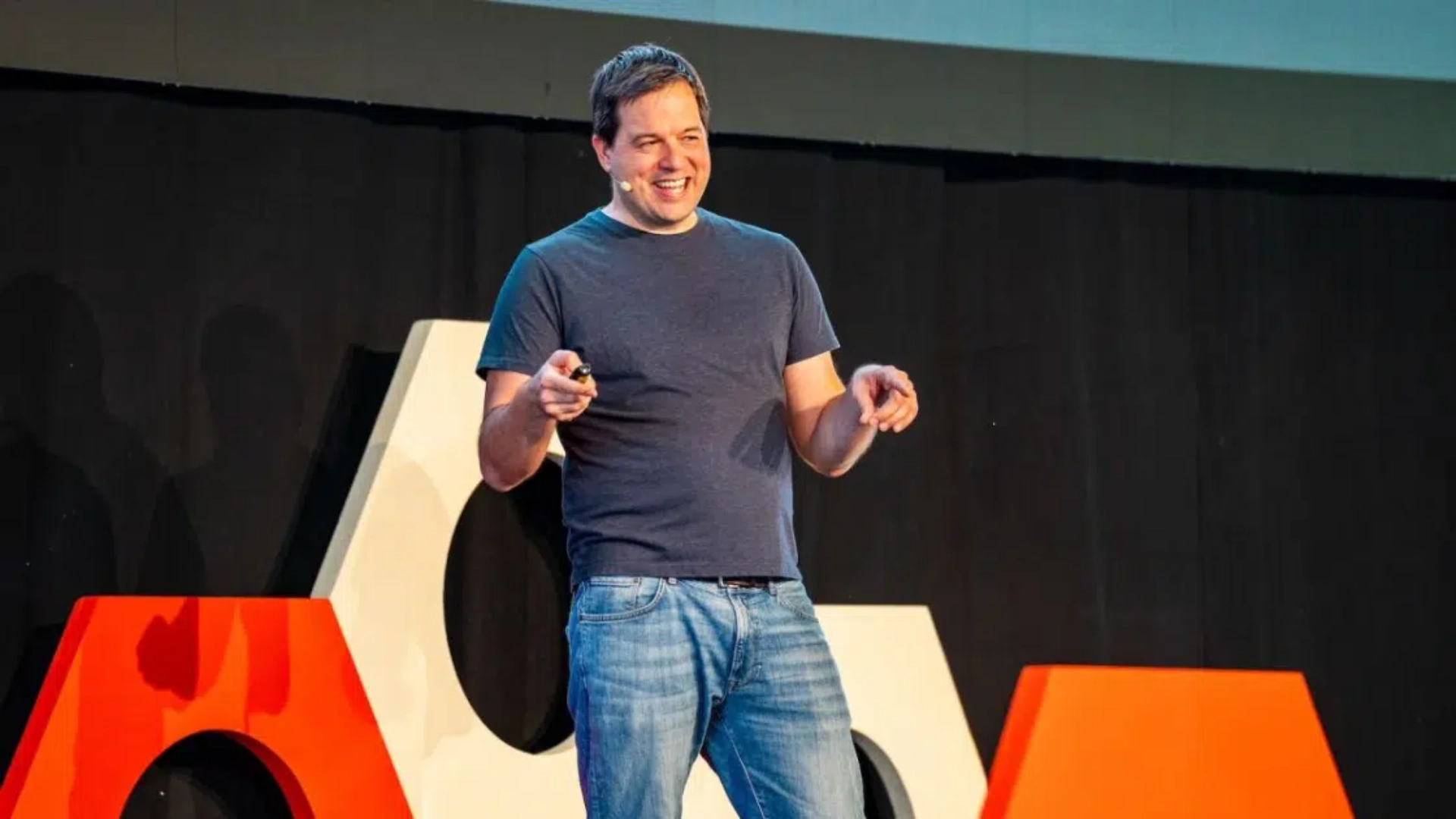 Gergely Orosz smiling on a conference stage with a headset microphone and presentation remote