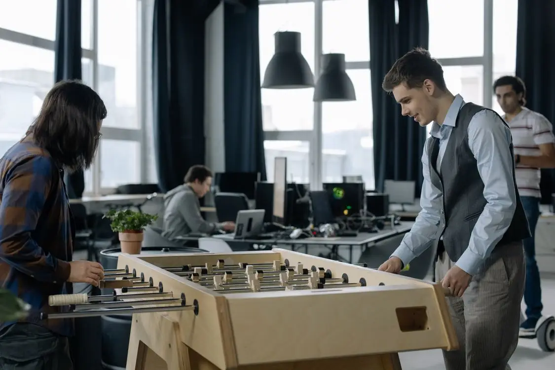 Two people playing foosball in a modern open-plan office space with large windows