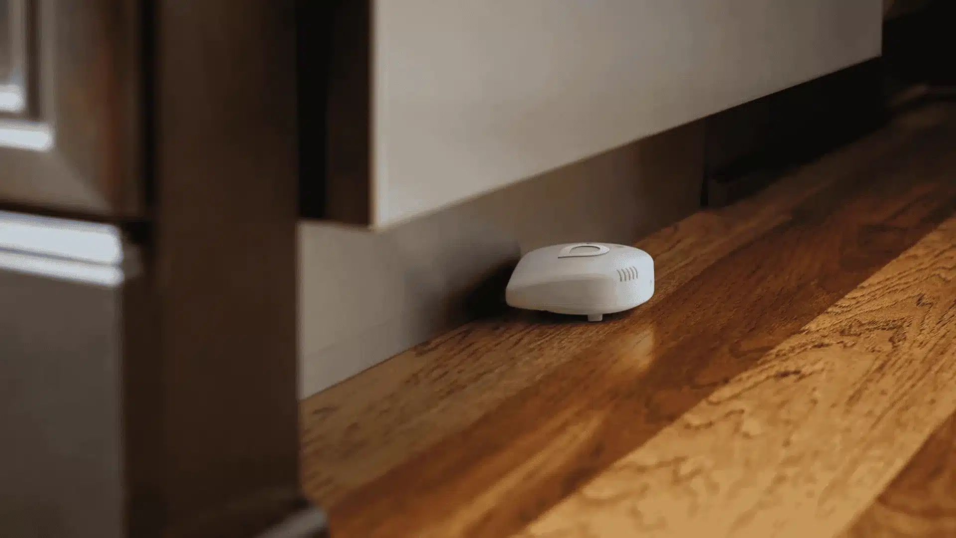 Moen Smart Leak Detector placed along a baseboard on hardwood flooring for early water leak detection in a home