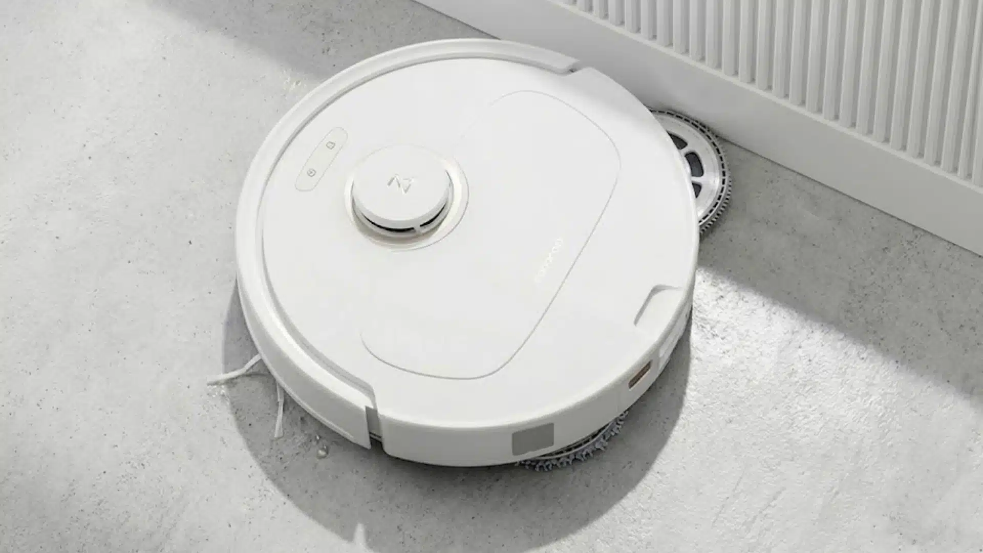 White Roborock robot vacuum mopping near a wall on a light floor with sunlight across the room