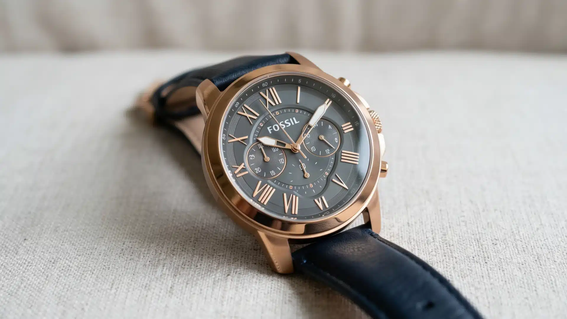 A close-up shot of a Fossil chronograph watch with a navy blue leather strap resting on a textured light beige surface