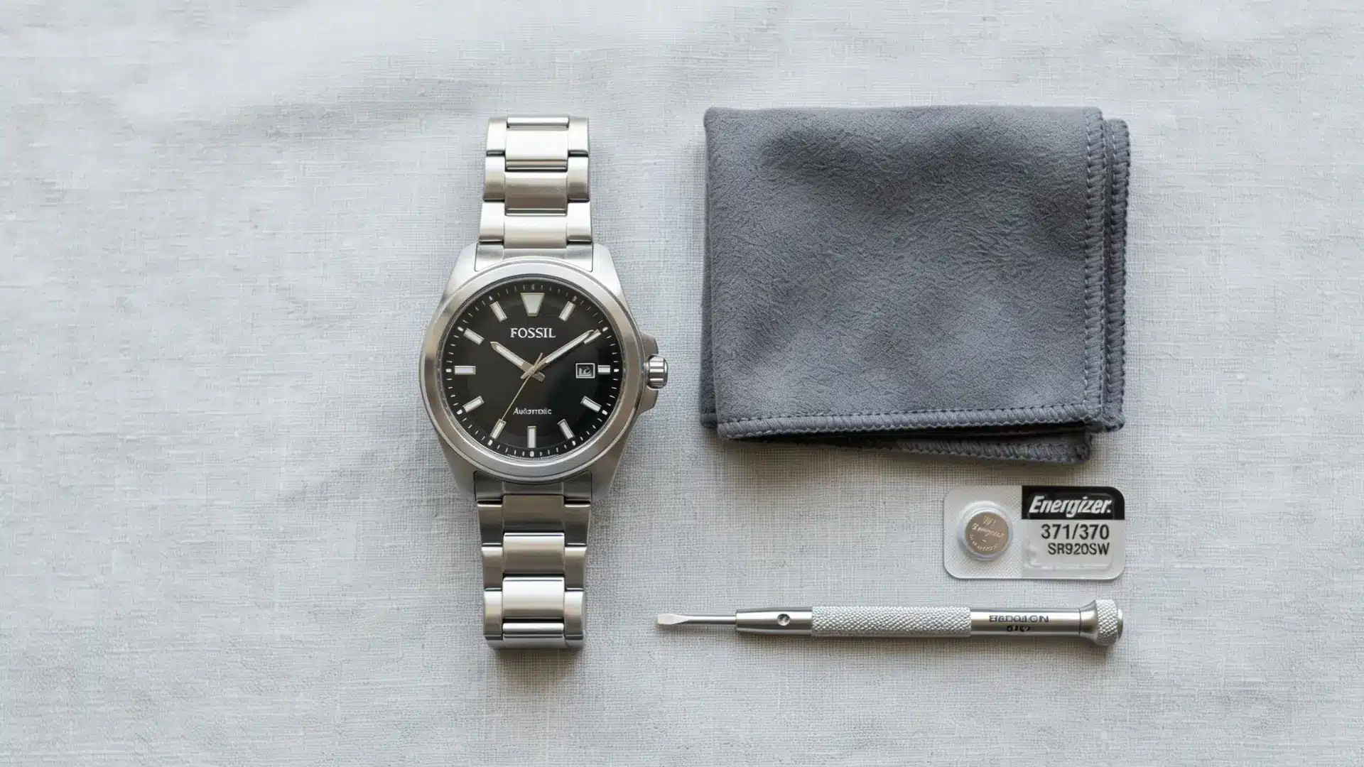 A Fossil watch with a black dial next to a cleaning cloth, a small screwdriver, and a watch battery