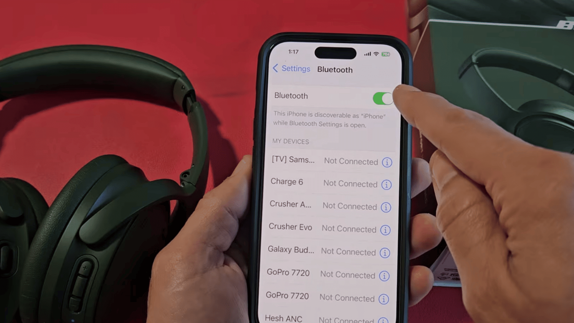a hand enabling bluetooth on an iphone while black over ear headphones rest beside the phone and retail box