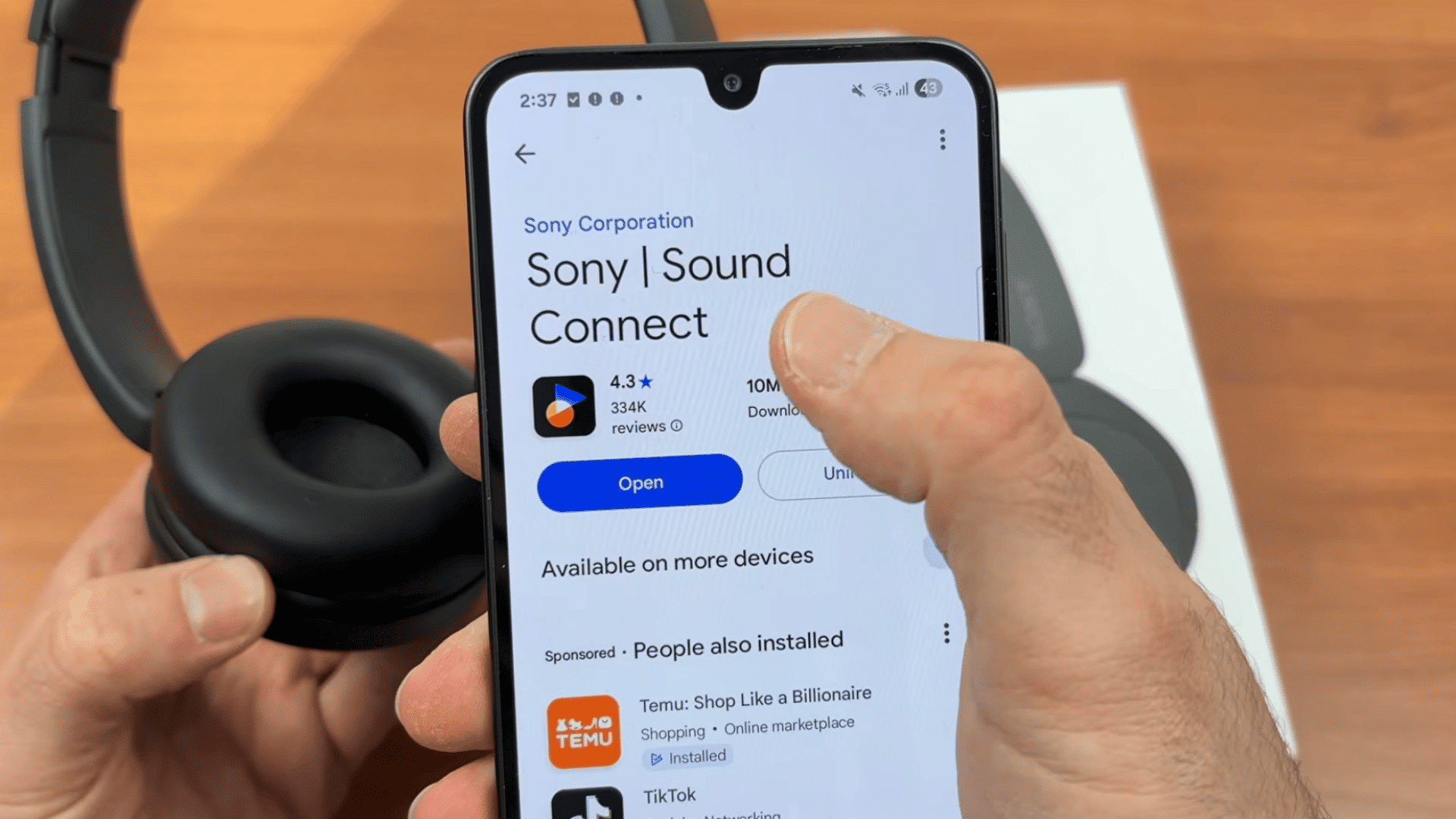 a hand opening the sony sound connect app on an android phone beside black over ear headphones and retail box