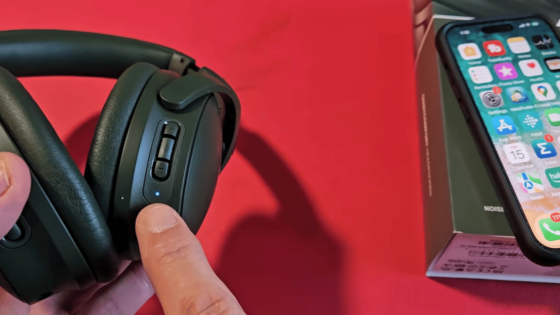 a hand pointing to the blue status light and control buttons on black over ear headphones beside an iphone