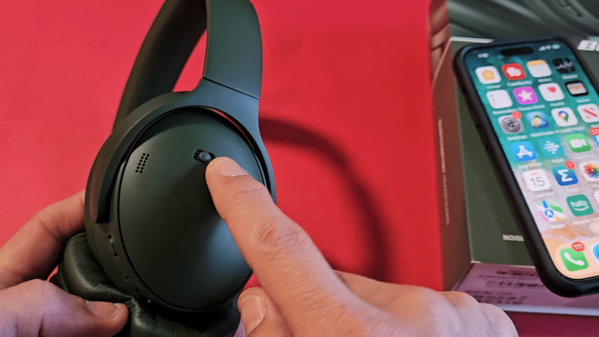 a hand pressing the bluetooth pairing button on black over ear headphones beside an iphone and product box