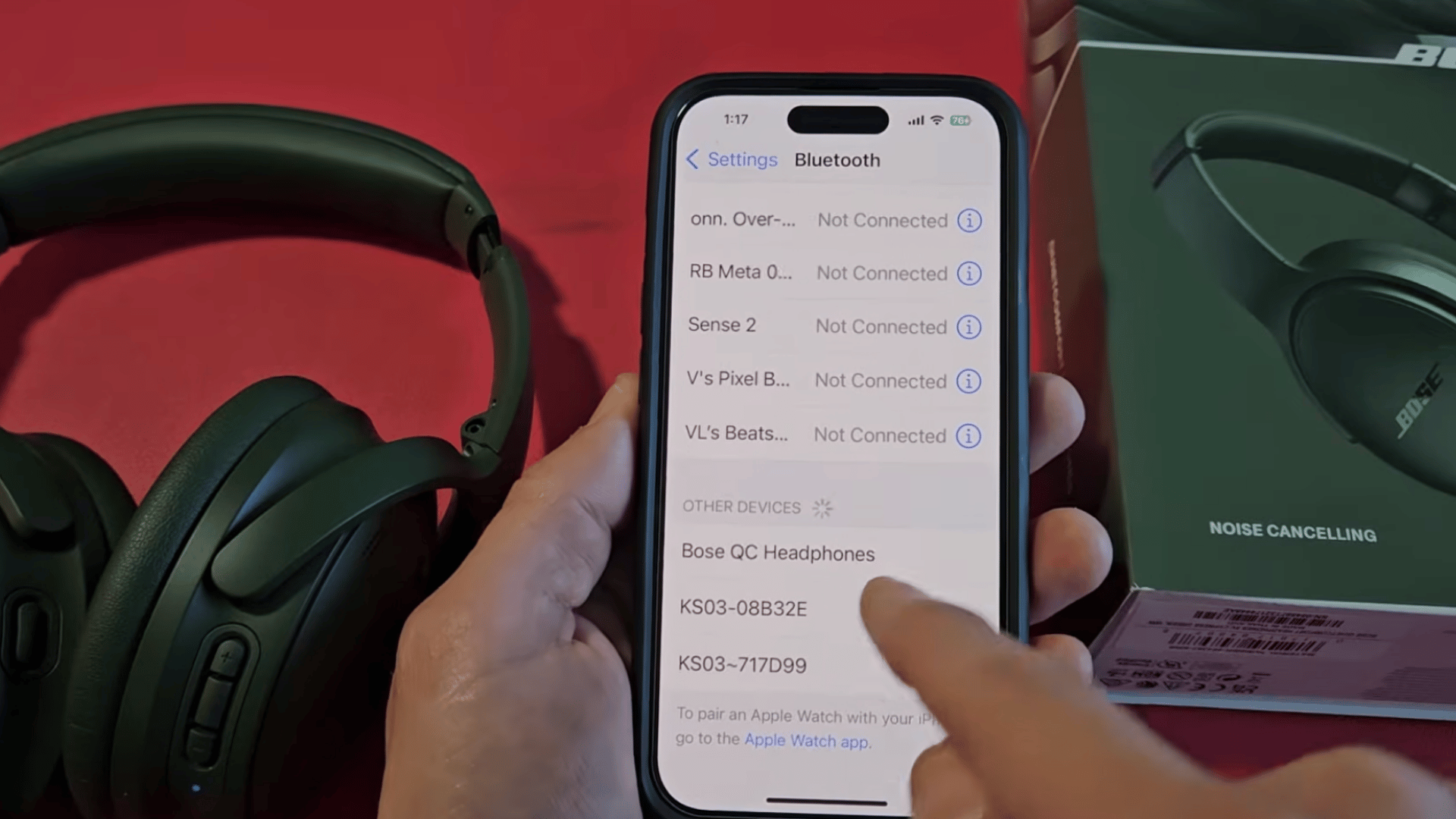 a hand selecting bose qc headphones from the available bluetooth devices list on an iphone beside the headphones