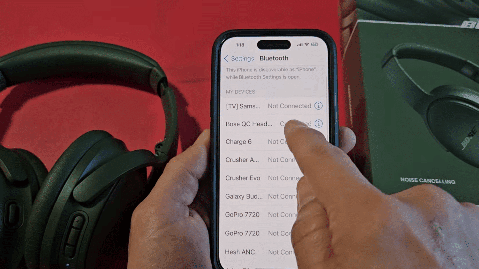 a hand tapping connected bose qc headphones in the iphone bluetooth settings beside the matching headphones box