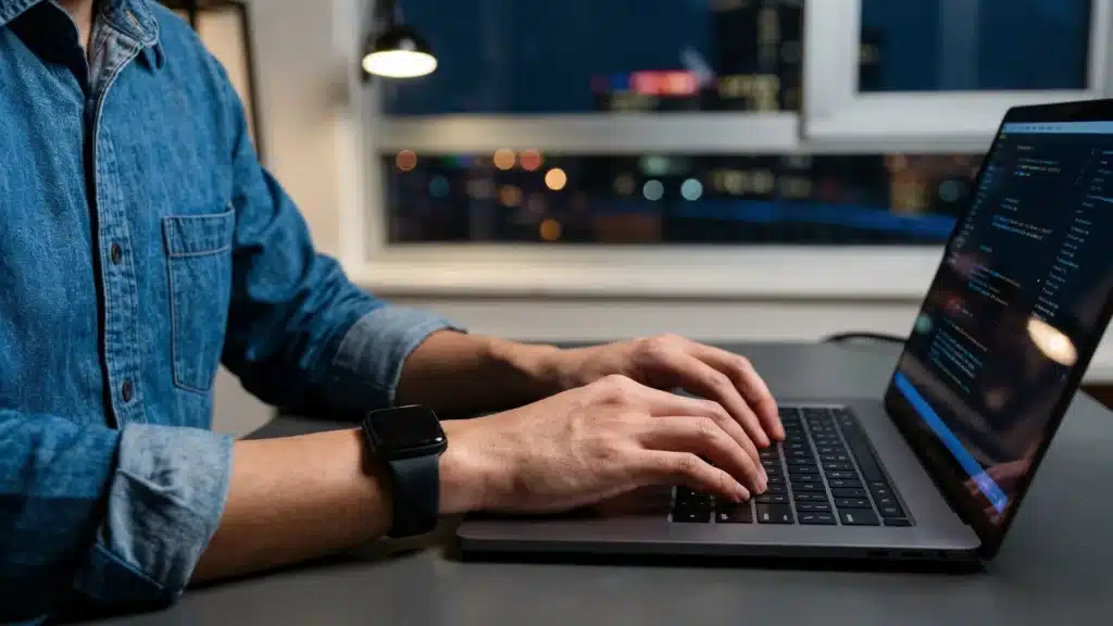 A programmer in a denim shirt types code on a laptop at night with blurry city lights in the background