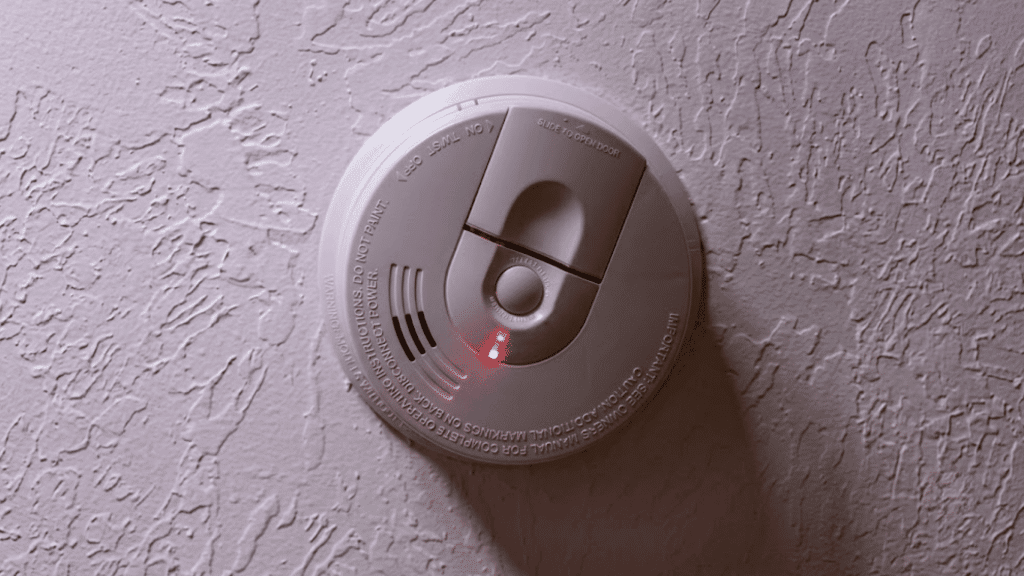 a white smoke detector mounted on a textured ceiling with a red indicator light glowing to show active status