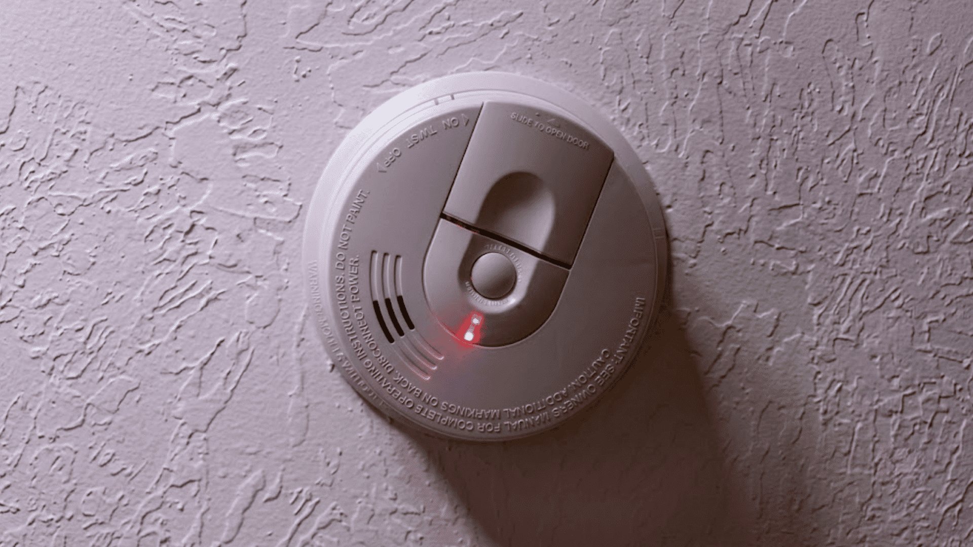 a white smoke detector mounted on a textured ceiling with a red indicator light glowing to show active status
