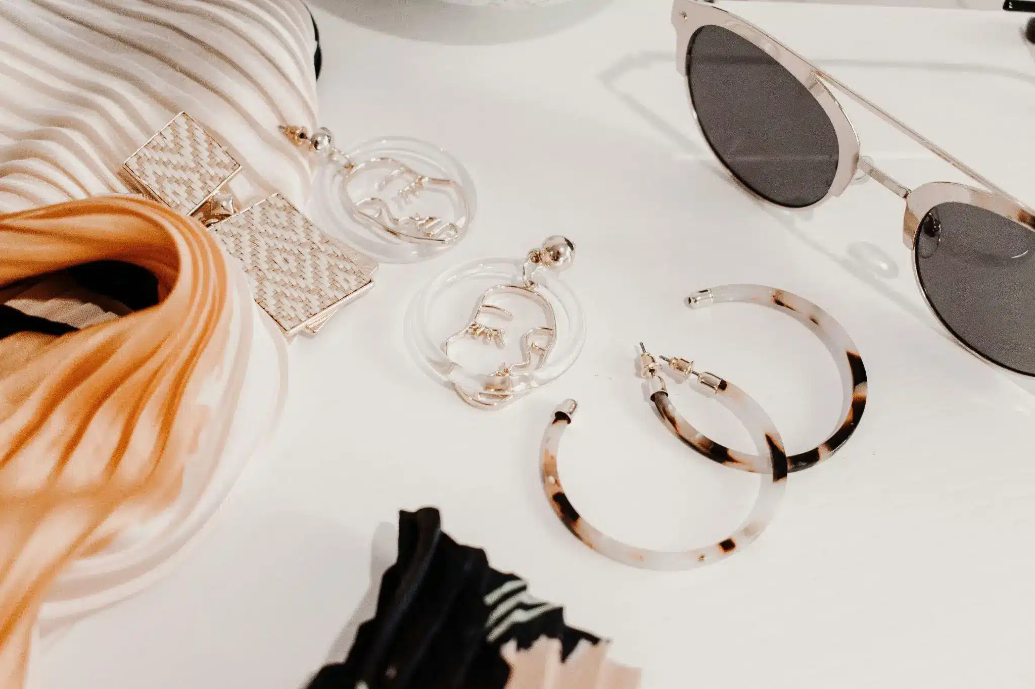 Stylish sunglasses and earrings arranged on white surface with patterned fabric and accessories