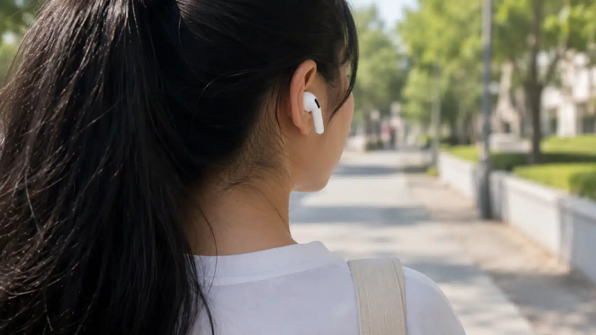 Black-haired woman wearing AirPods on a sunny outdoor walkway from a rear side view