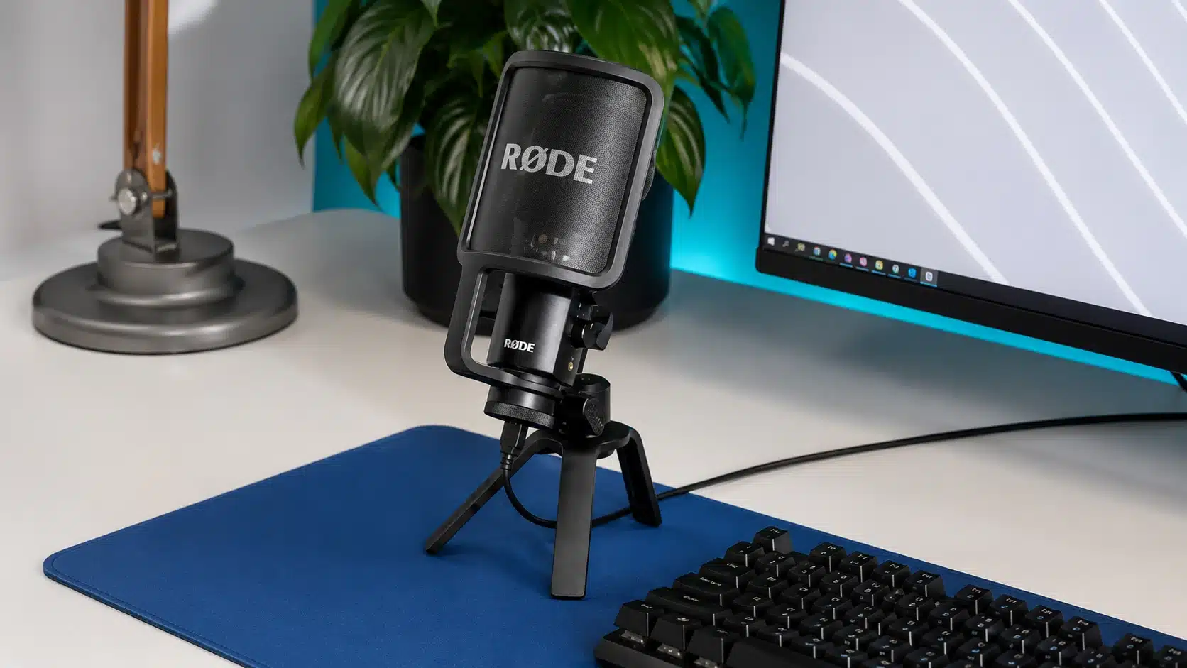 black røde desktop microphone on tripod over blue desk mat beside keyboard, monitor, plant, and modern workspace