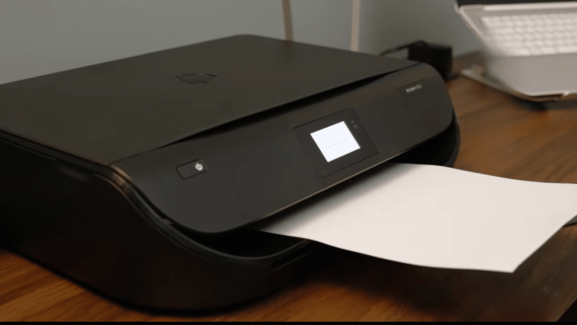 black wireless printer producing a blank sheet on a wooden desk beside an open laptop in a home office
