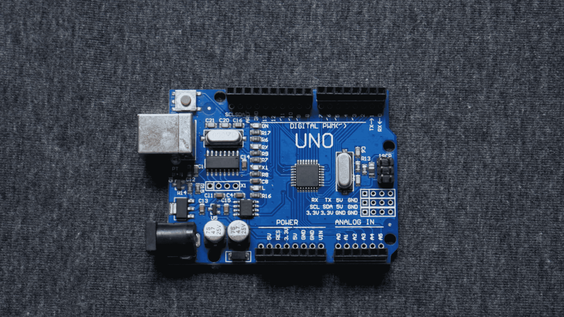 blue arduino uno microcontroller board with usb port and pin headers placed on a dark fabric background