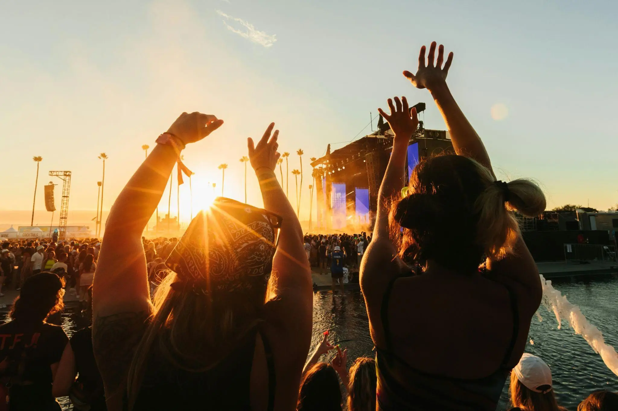 Crowd raising hands at outdoor concert during sunset, with stage and palm trees in background