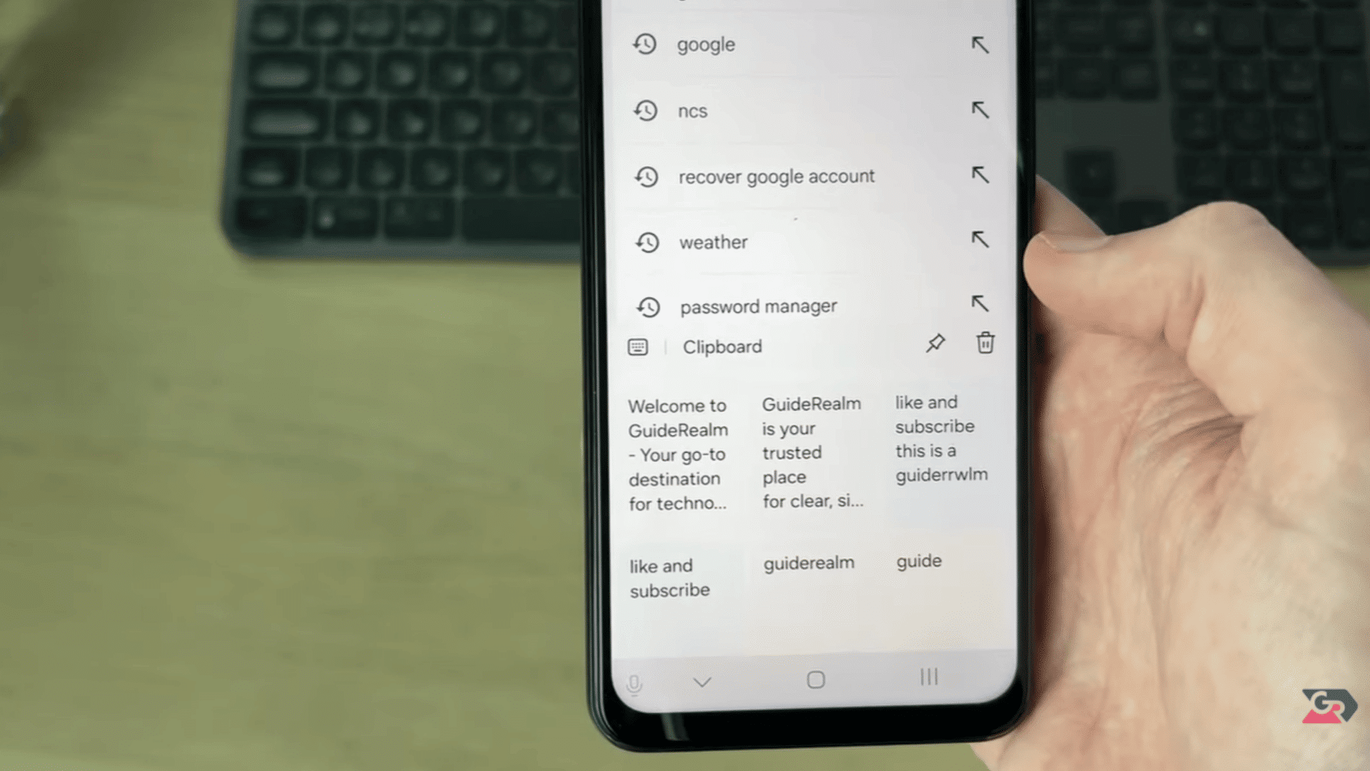 hand holding a smartphone displaying clipboard history and recent searches with a desktop keyboard in the background