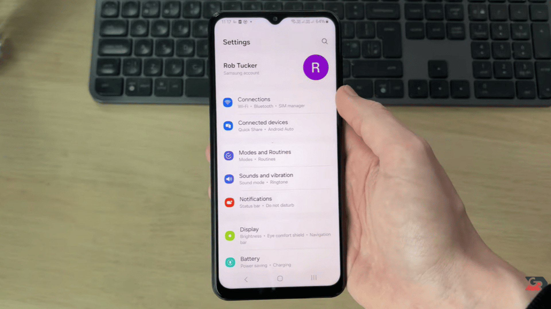 hand holding a smartphone displaying the settings menu with a wireless keyboard on a desk in the background