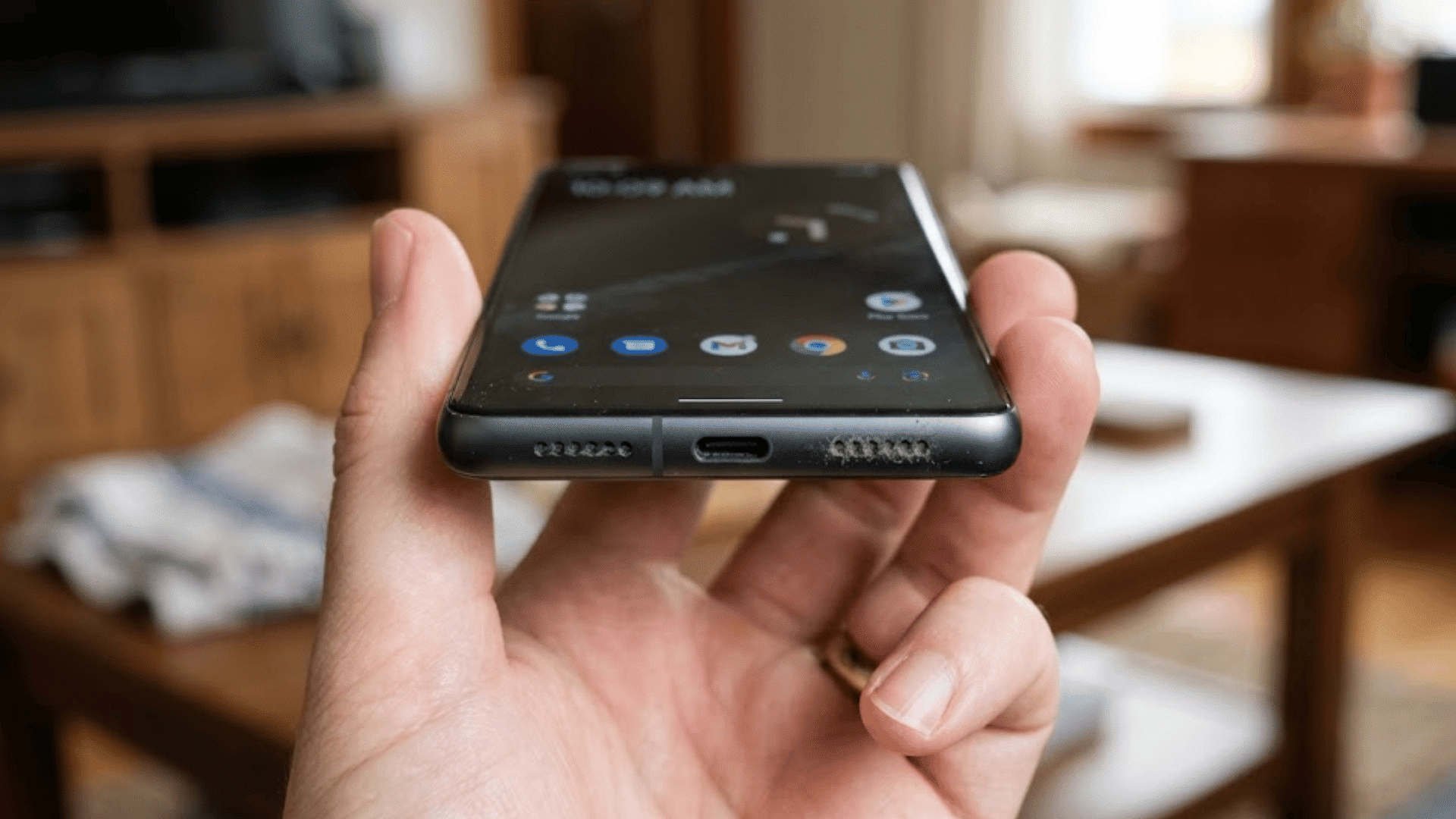 hand holding a smartphone showing dust buildup in the bottom speaker grilles beside the charging port inside a home setting