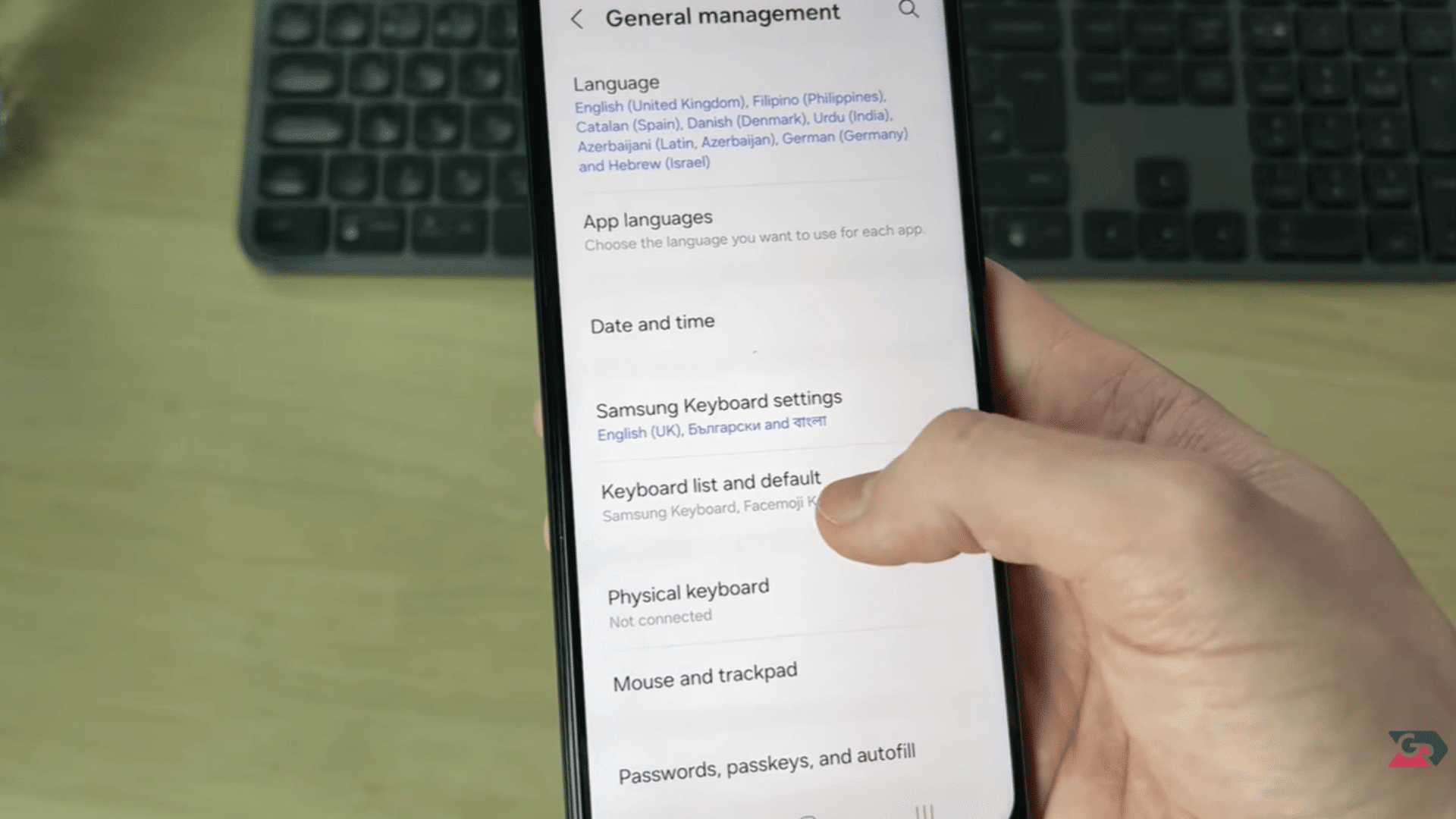hand using a smartphone showing general management settings with keyboard options and a desktop keyboard behind
