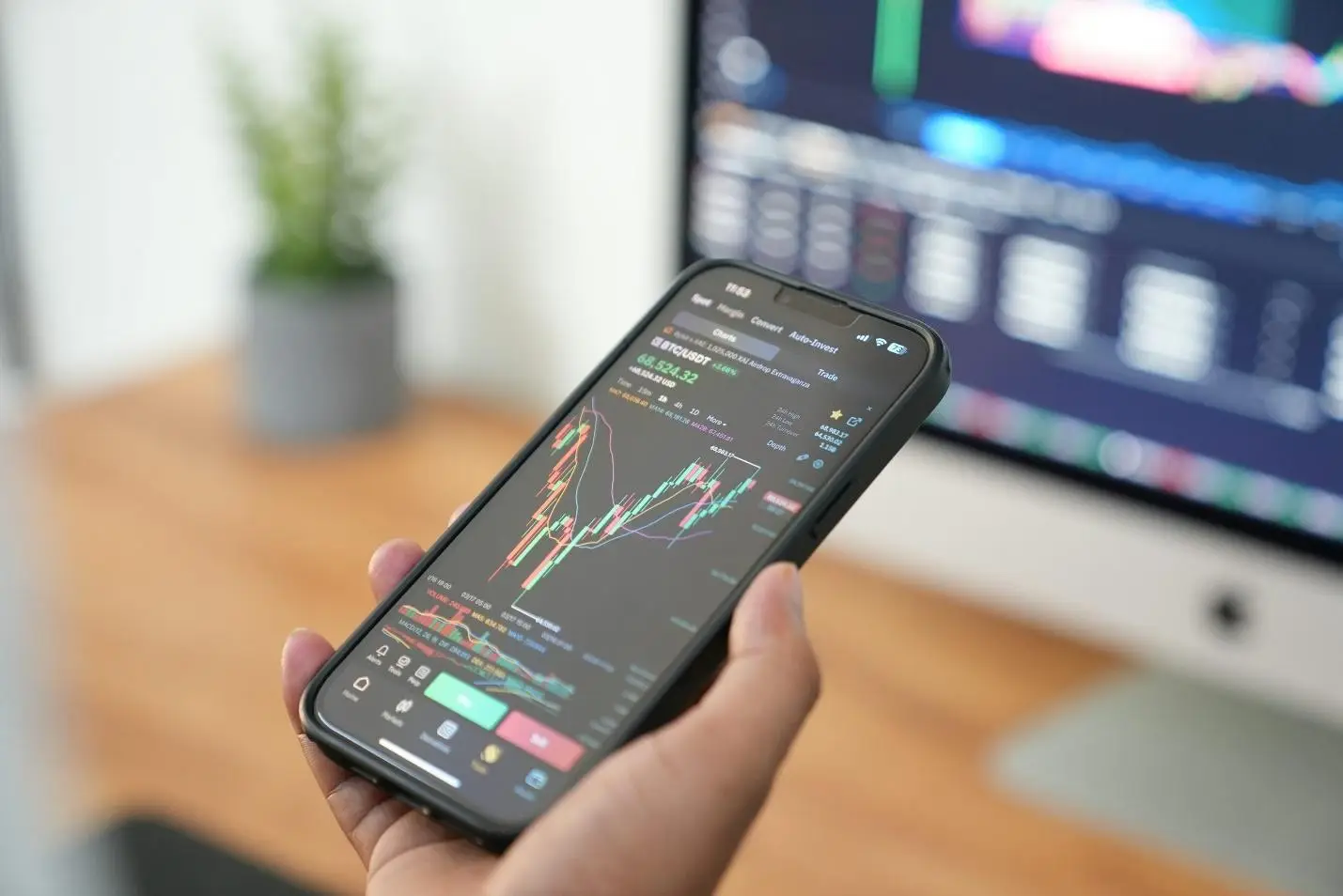 Hand holding smartphone displaying stock market charts in bright office setting