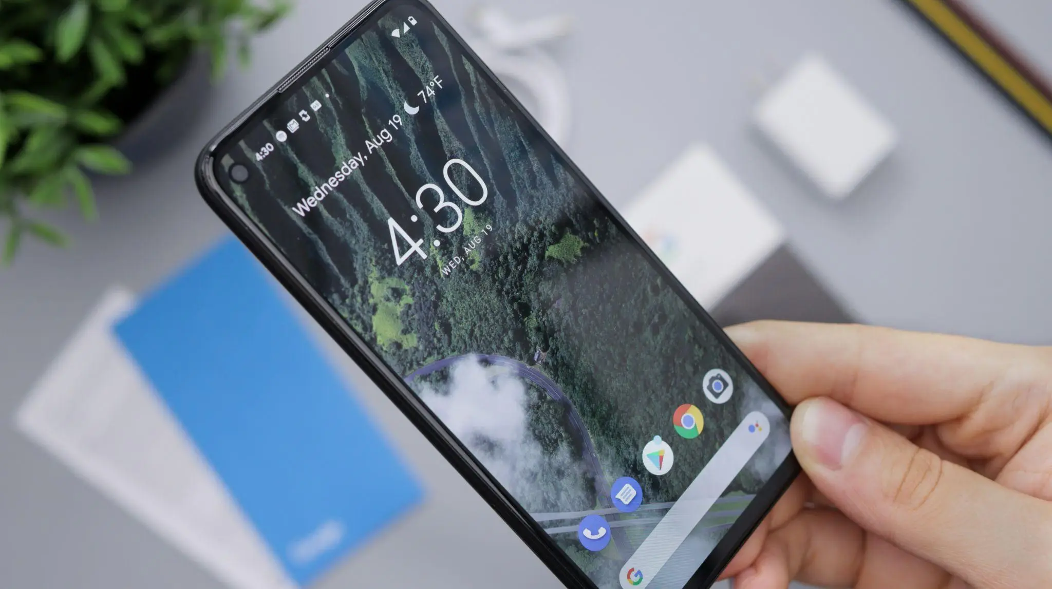 Person holding smartphone displaying forest-themed wallpaper on a table with tech items nearby