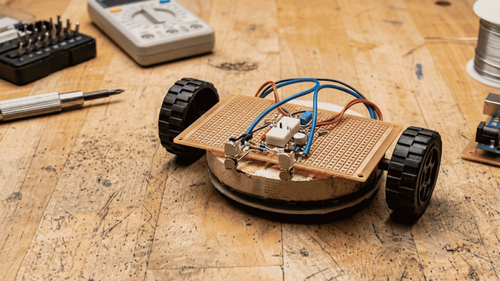 simple homemade robot with exposed circuit board wires and two wheels moving across a wooden surface