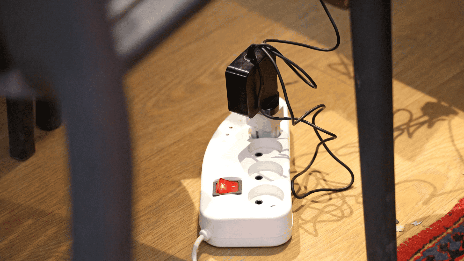white power strip with connected charger and tangled cables placed on a wooden floor beneath a table