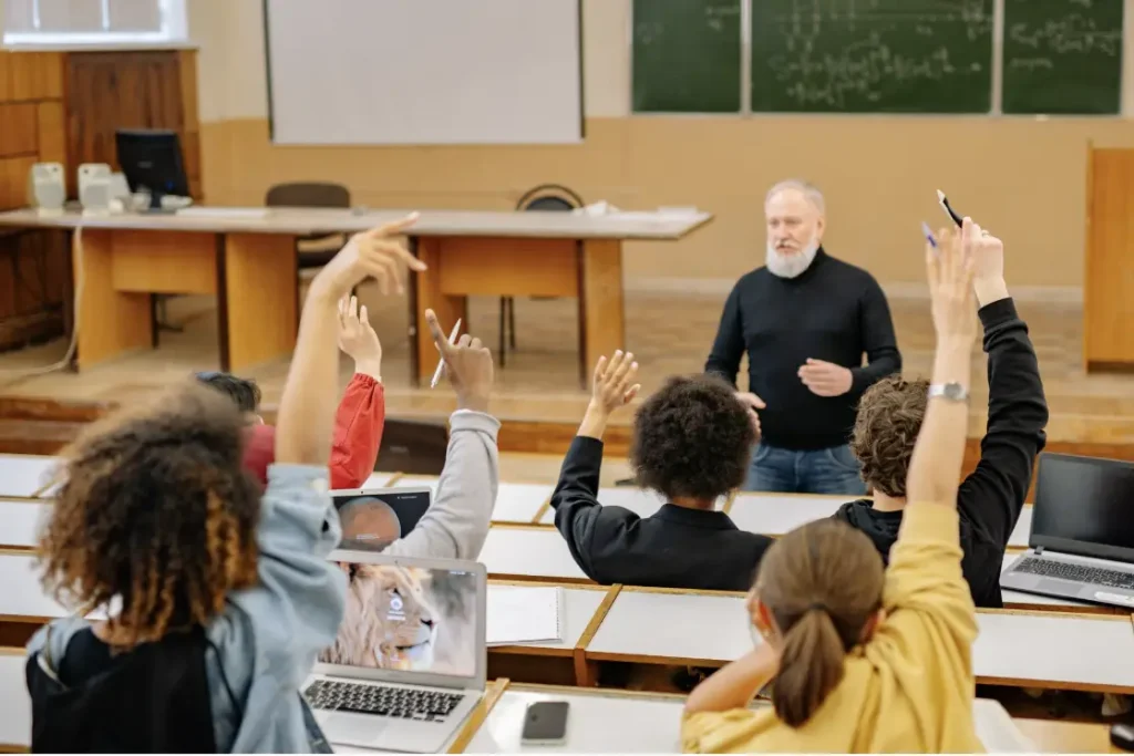 Students raising hands in classroom during lecture with bearded professor at front, laptops on desks
