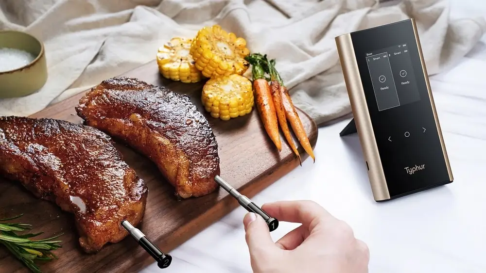 Grilled steaks with corn and carrots on wooden board beside digital cooking thermometer