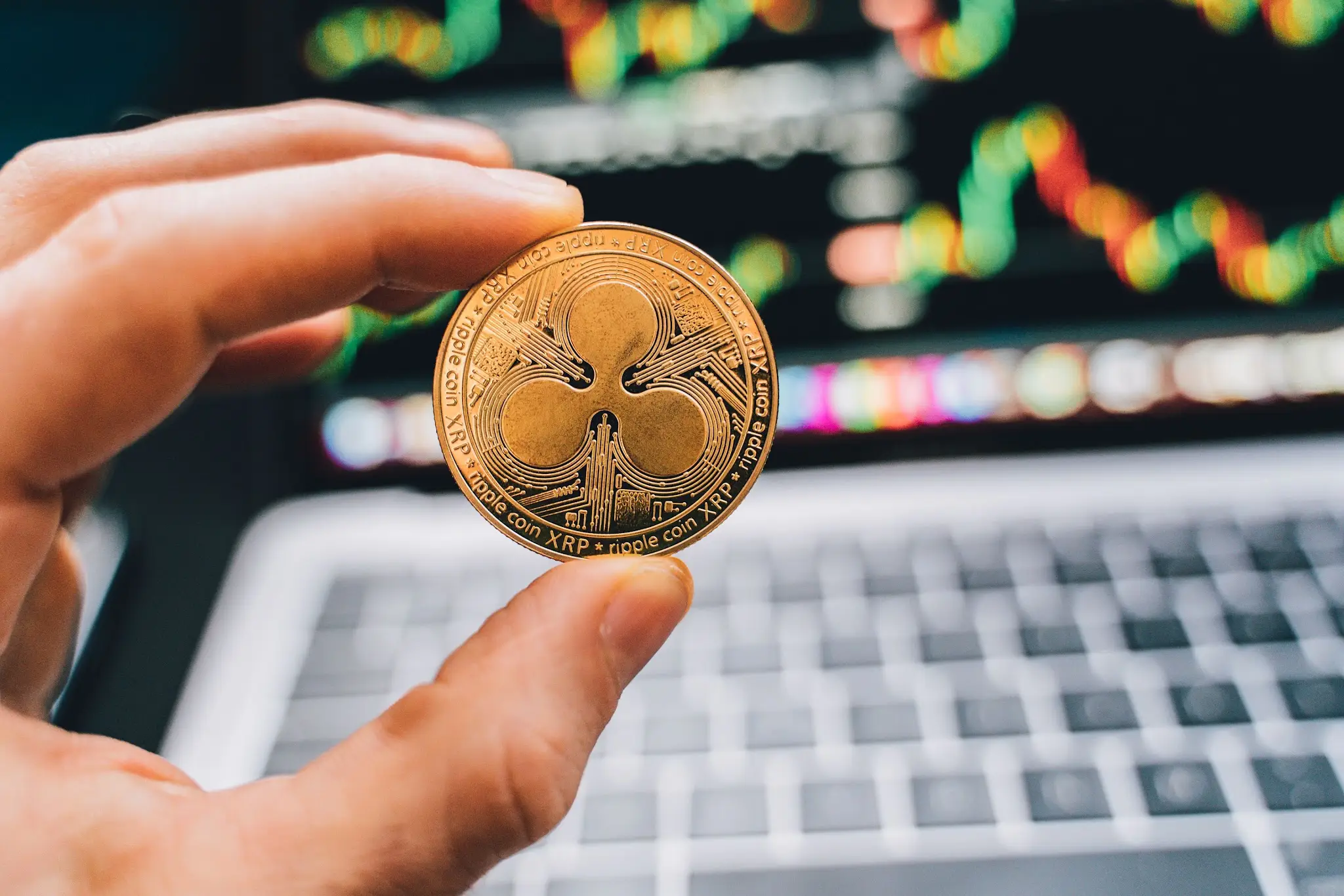 Hand holding a Ripple coin in front of a laptop displaying cryptocurrency charts