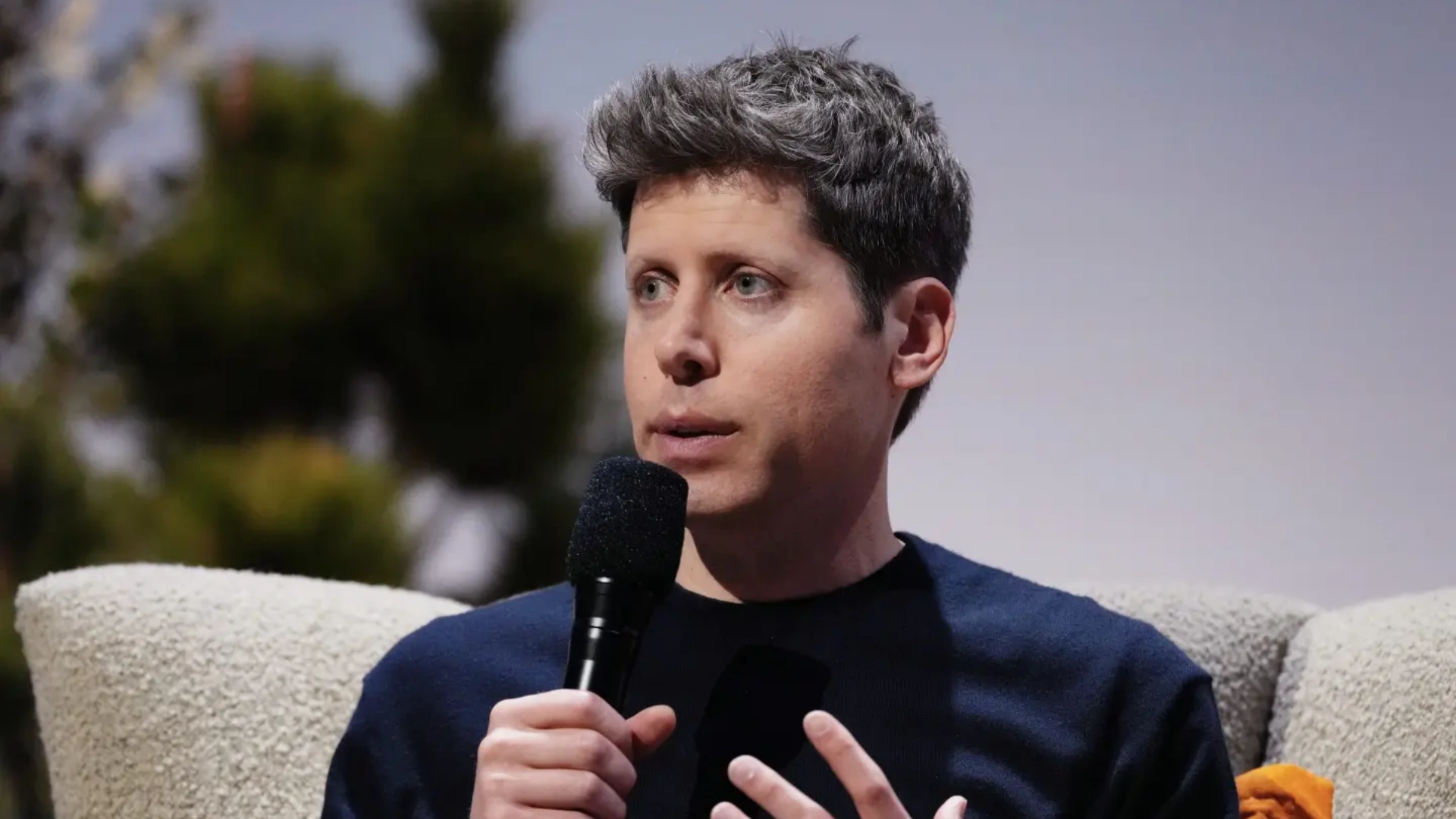 sam altman speaking into a microphone during an outdoor interview with a blurred background and wearing a dark sweater